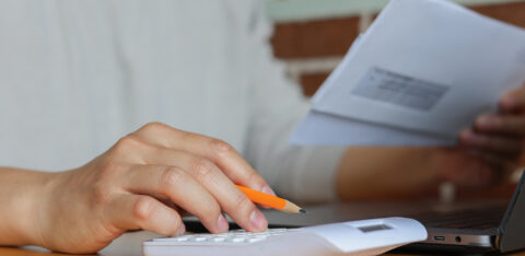 Close-up of hands using a white calculator and holding an envelope, with a pencil and laptop on a wooden desk.