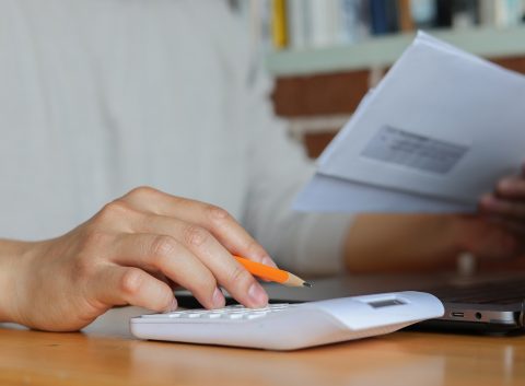 Close-up of hands using a white calculator and holding an envelope, with a pencil and laptop on a wooden desk.