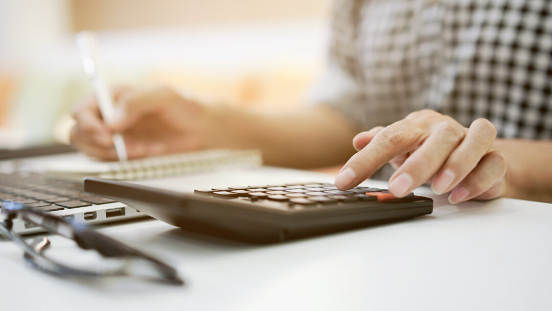 Close-up of a person using a calculator while taking notes next to a laptop on a desk.