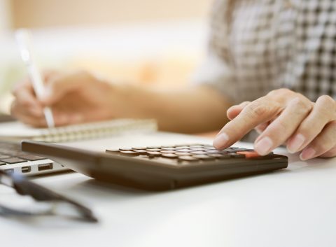 Close-up of a person using a calculator while taking notes next to a laptop on a desk.