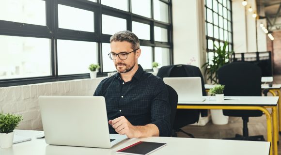 Focused man working on a laptop at a modern office desk with large windows and natural light.