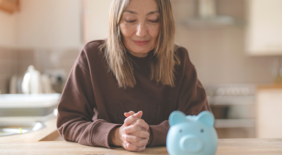 Middle-aged woman sitting at a kitchen table, looking thoughtfully at a blue piggy bank and calculator in front of her.