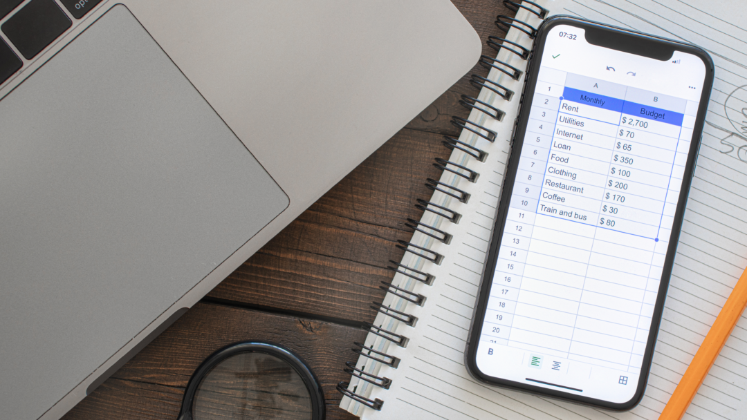Smartphone displaying a monthly budget spreadsheet next to a laptop and notebook