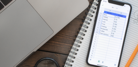 Smartphone displaying a monthly budget spreadsheet next to a laptop and notebook