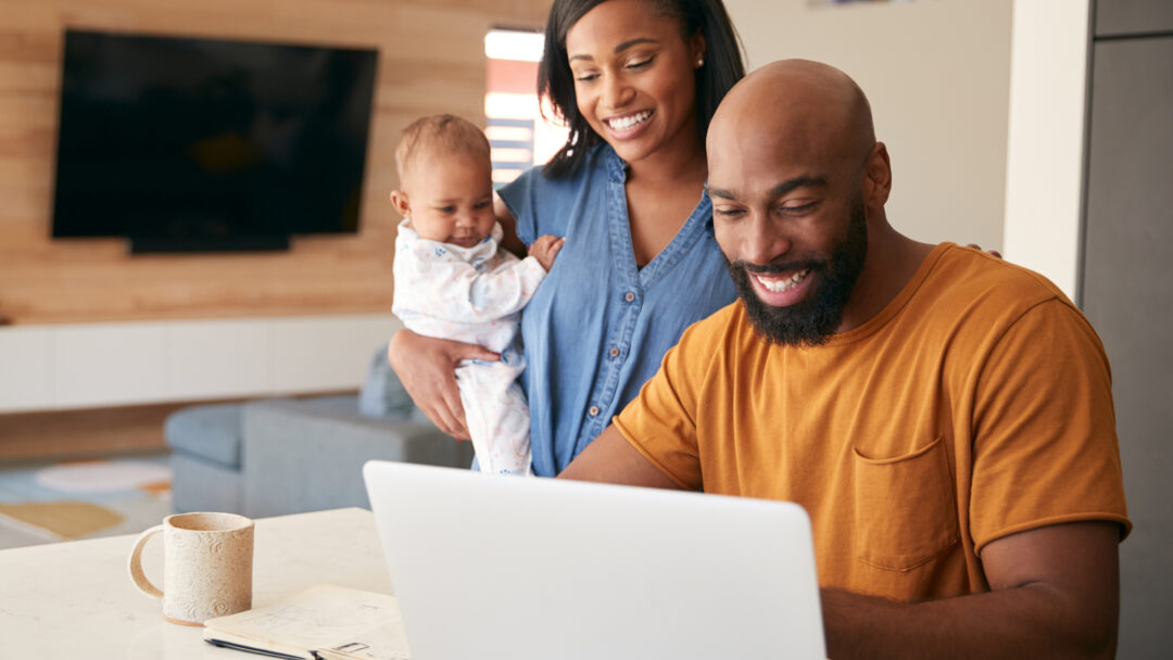 Smiling couple with a baby looking at a laptop together at home, appearing happy and engaged.