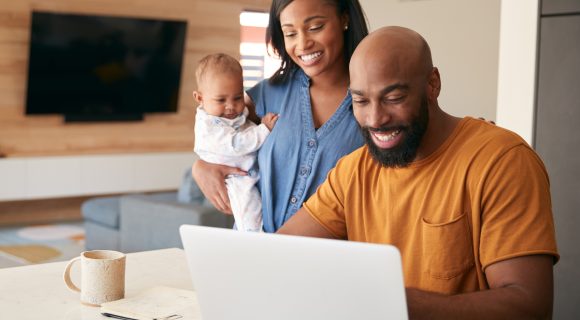 Smiling couple with a baby looking at a laptop together at home, appearing happy and engaged.