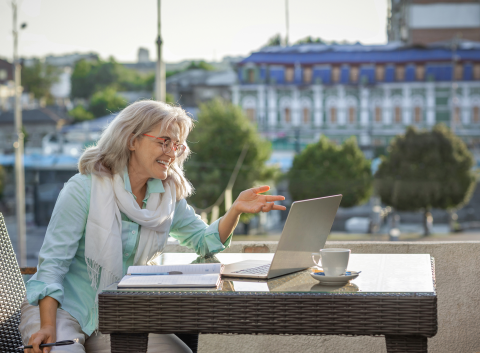 A smiling older woman sits at an outdoor café table, having a video call on her laptop with a notebook and coffee beside her.