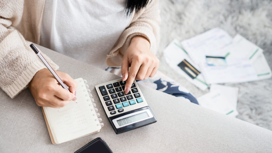 Woman calculating expenses with a large calculator and taking notes in a notebook, surrounded by bills and a smartphone.