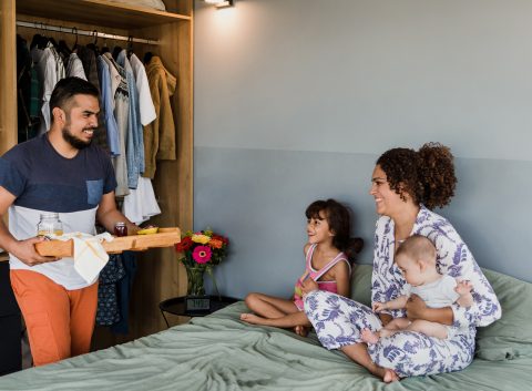 A father brings breakfast in bed to a smiling mother and their two children, celebrating a relaxed morning at home.