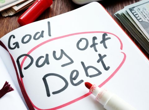 Notebook page with the words “Goal: Pay Off Debt” circled in red marker, surrounded by cash and a red pen.