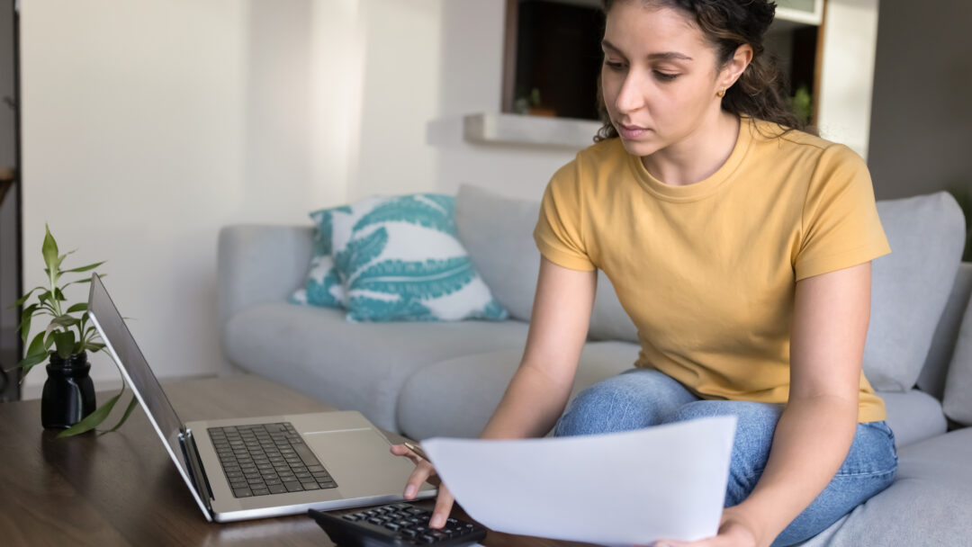 Young woman sitting on a couch, reviewing financial documents with a calculator and laptop on the coffee table.