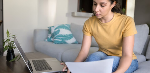 Young woman sitting on a couch, reviewing financial documents with a calculator and laptop on the coffee table.