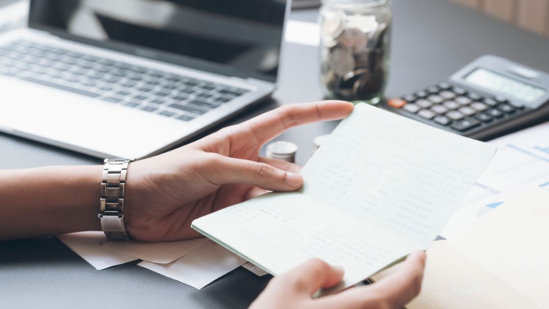 Person reviewing a bank book at a desk with a laptop, calculator, and jar of coins
