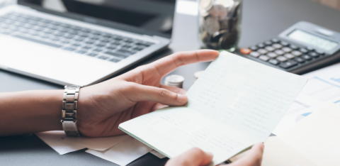 Person reviewing a bank book at a desk with a laptop, calculator, and jar of coins