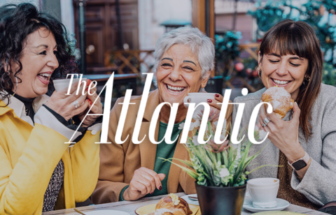 Lively café scene with three women chatting and smiling, holding coffee cups and pastries, branded with The Atlantic logo.