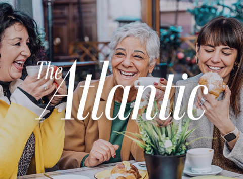Lively café scene with three women chatting and smiling, holding coffee cups and pastries, branded with The Atlantic logo.