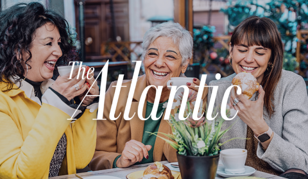 Lively café scene with three women chatting and smiling, holding coffee cups and pastries, branded with The Atlantic logo.