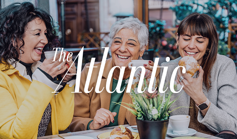 Lively café scene with three women chatting and smiling, holding coffee cups and pastries, branded with The Atlantic logo.