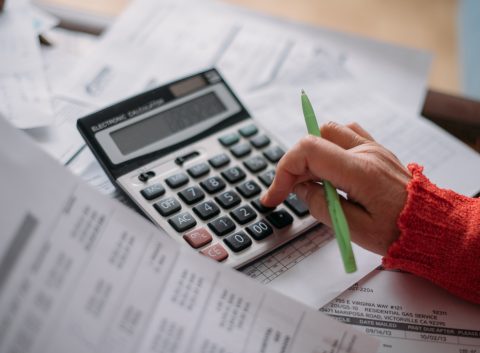 Close-up of a person using a calculator and reviewing bills, surrounded by financial documents and holding a green pen.