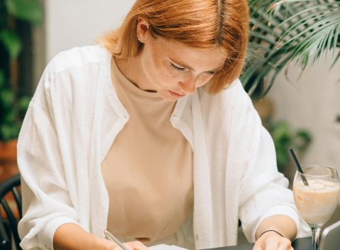 Woman focused on filling out financial documents at a desk with a laptop, calculator, magnifying glass, and iced coffee.