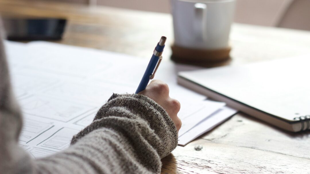 Close-up of a person writing on paper at a wooden desk with a notebook and coffee mug nearby.